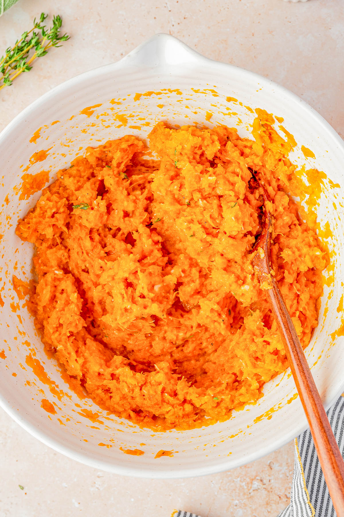 A white mixing bowl filled with a mashed orange mixture being stirred with a wooden spoon.
