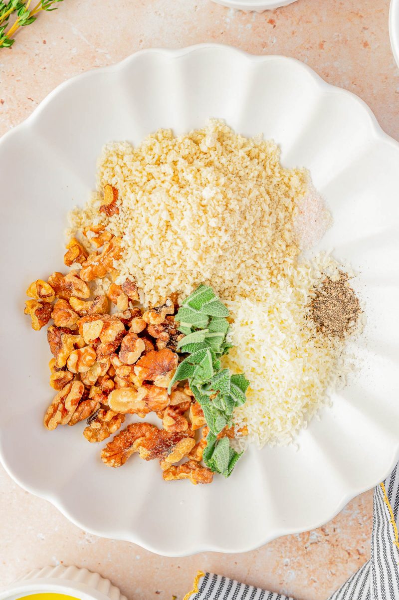 White plate with piles of panko breadcrumbs, chopped walnuts, grated cheese, black pepper, fresh sage leaves, and salt on a light countertop.