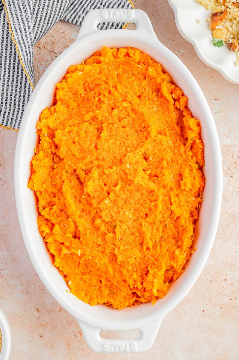 Oval white dish filled with mashed sweet potatoes on a light countertop, with part of a cloth napkin and another dish partially visible nearby.