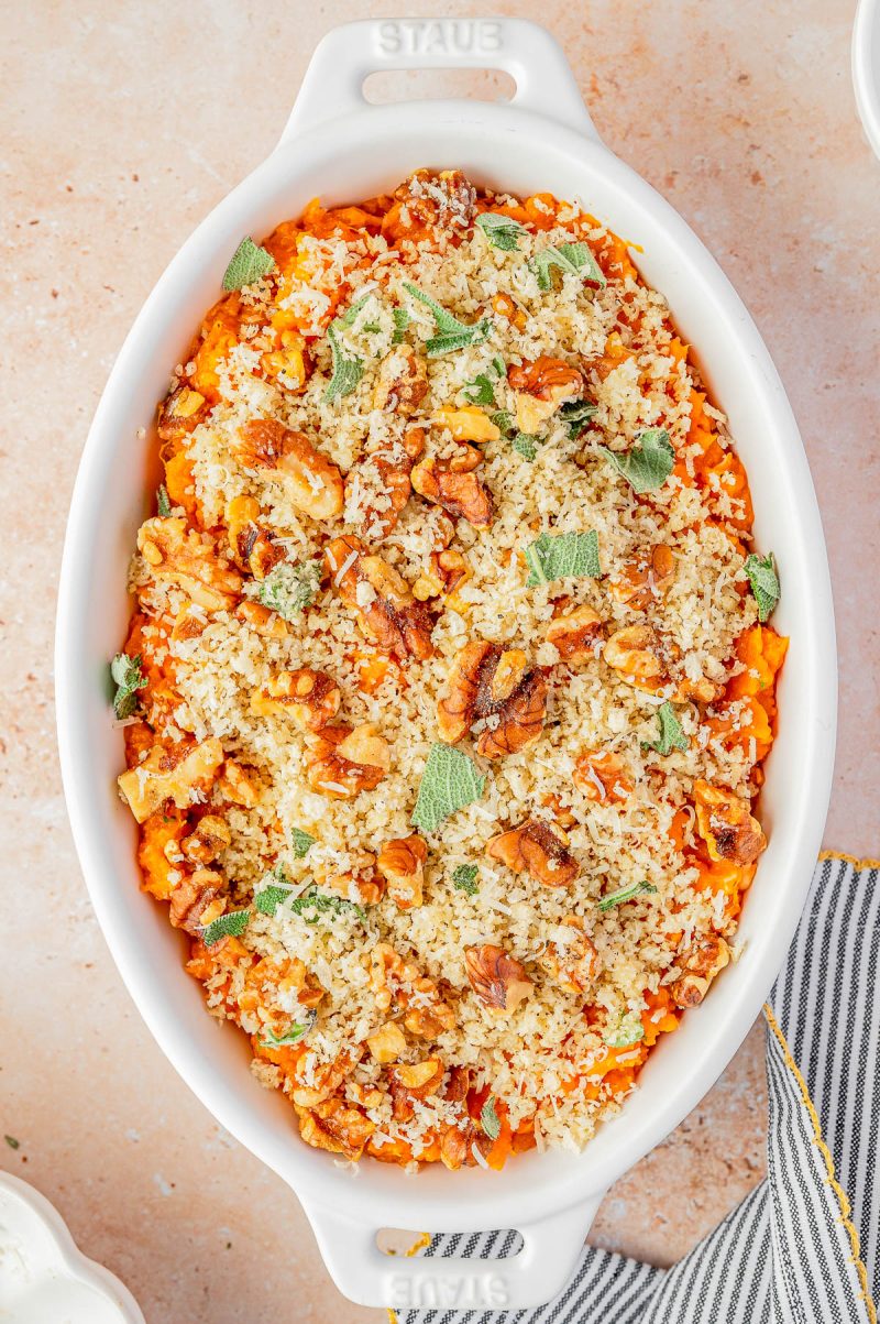 A white oval baking dish filled with a baked casserole topped with breadcrumbs, chopped nuts, and fresh herbs on a light surface.