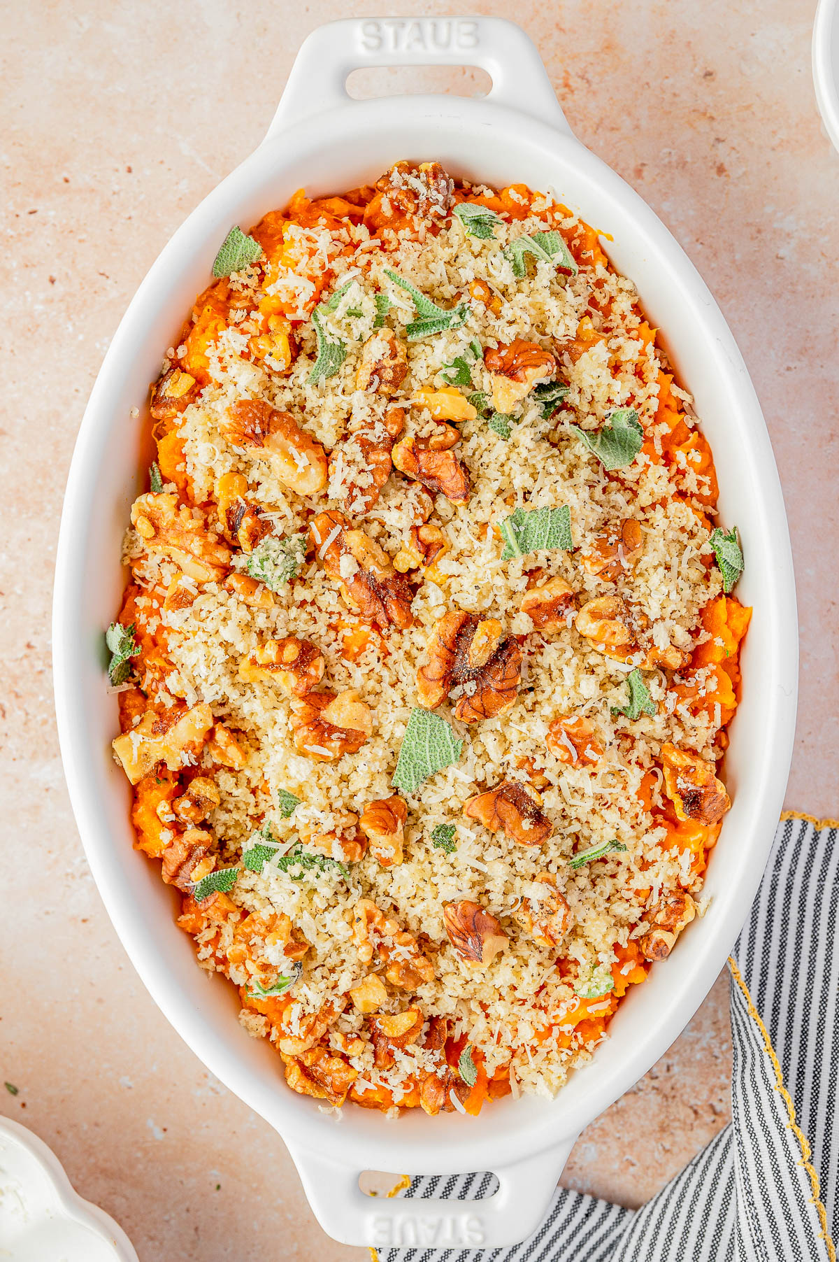 A white oval baking dish filled with a baked casserole topped with breadcrumbs, chopped nuts, and fresh herbs on a light surface.