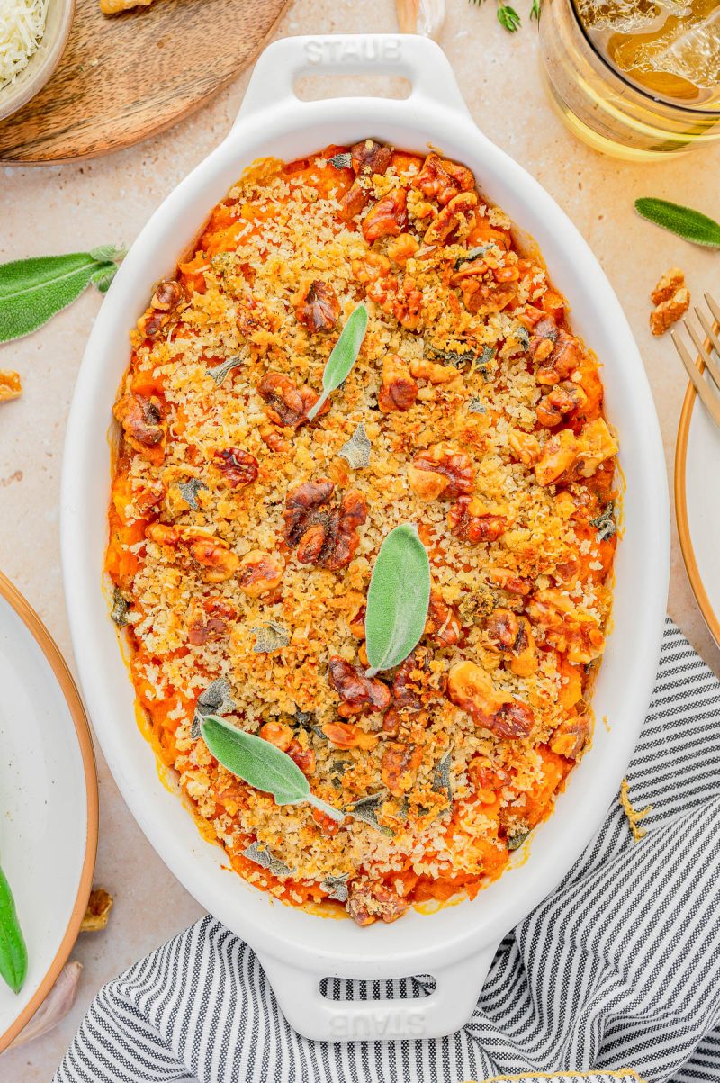 A baked casserole topped with breadcrumbs, walnuts, and sage leaves is presented in a white oval dish.
