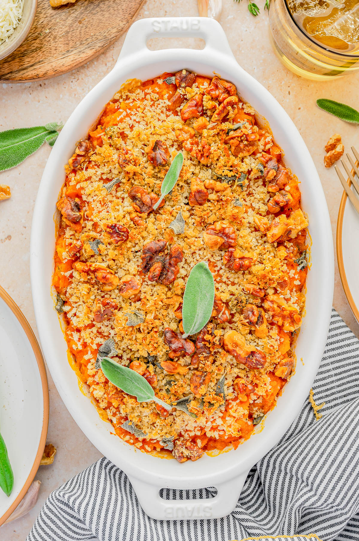 A baked casserole topped with breadcrumbs, walnuts, and sage leaves is presented in a white oval dish.
