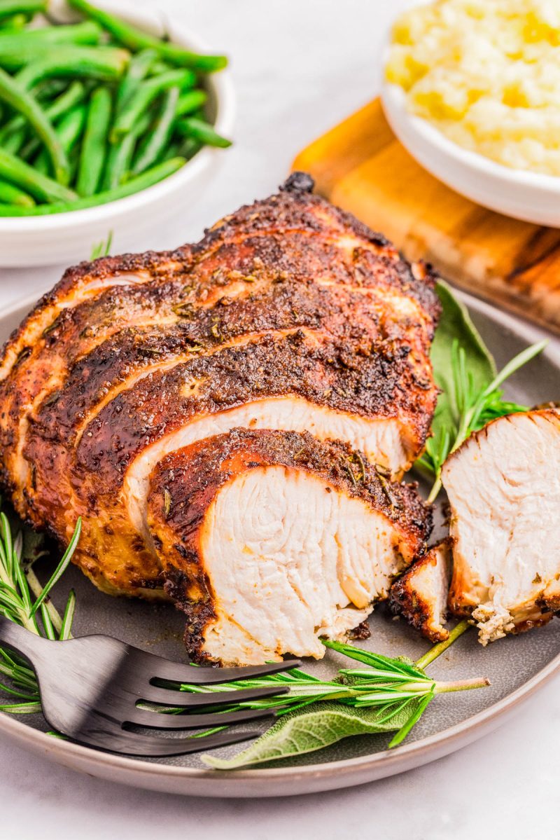Sliced roasted turkey breast served on a plate with fresh rosemary, accompanied by bowls of green beans and mashed potatoes in the background.