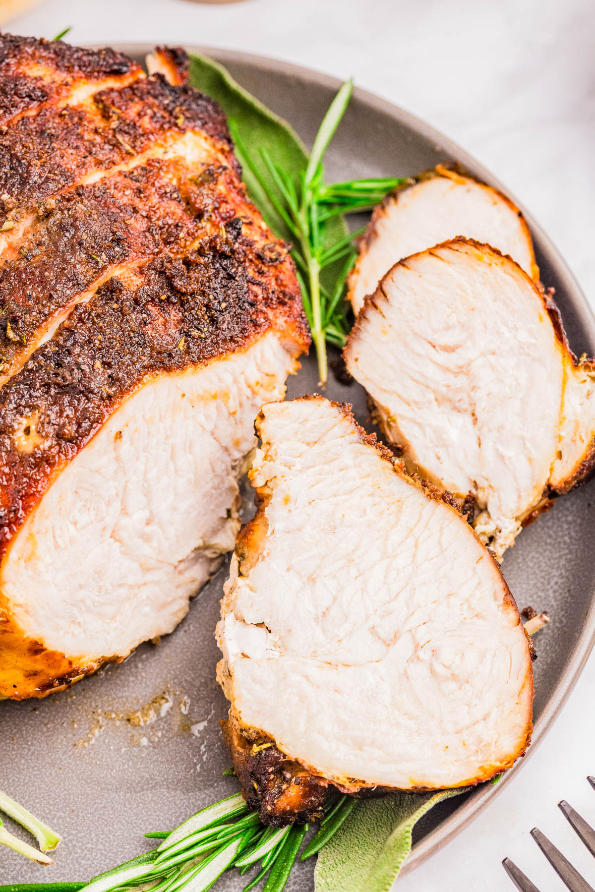 A plate with a roasted, seasoned pork loin cut into thick slices, garnished with fresh rosemary and sage.