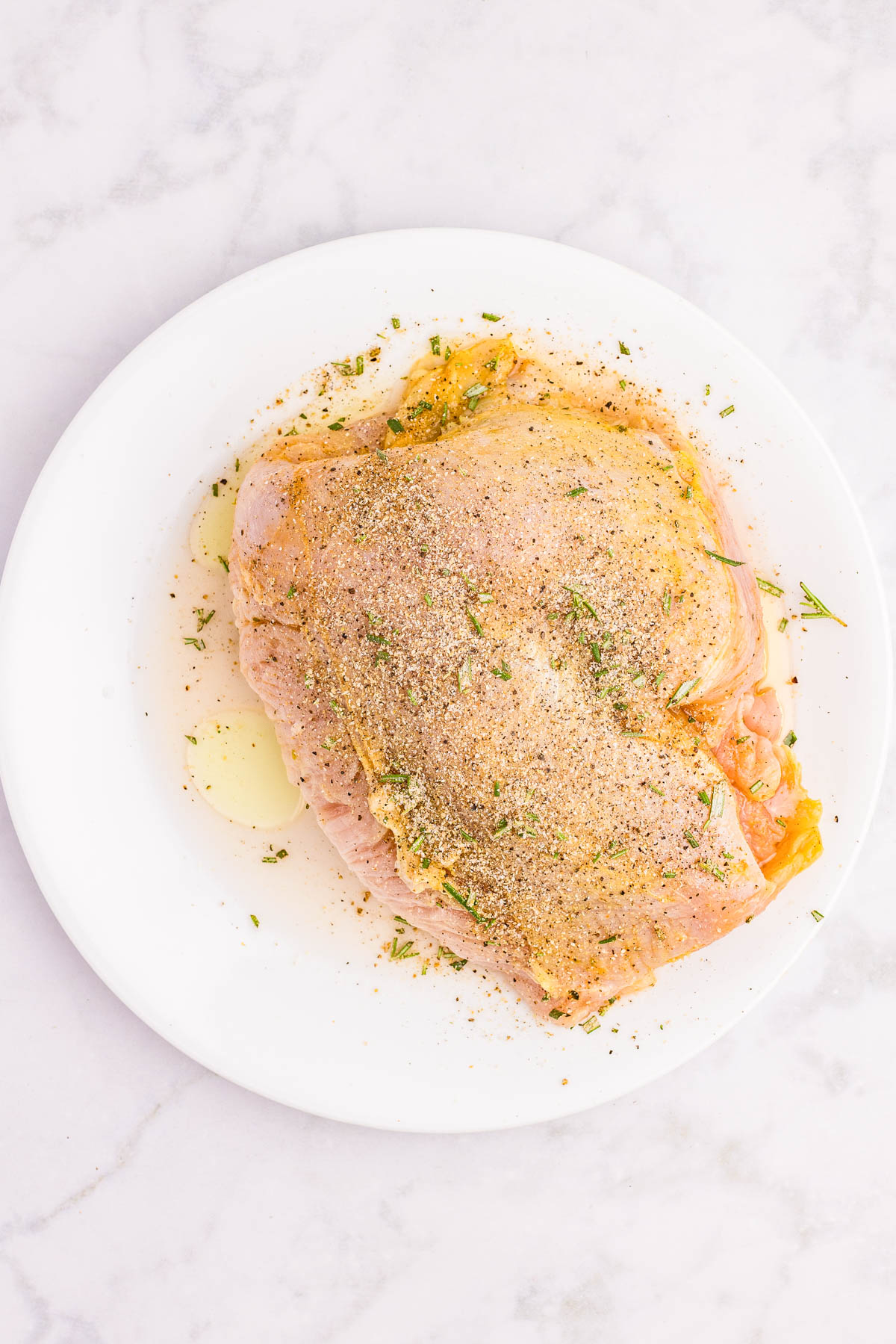 Raw turkey breast on a white plate, seasoned with olive oil, pepper, herbs, and spices, set on a light marble surface.