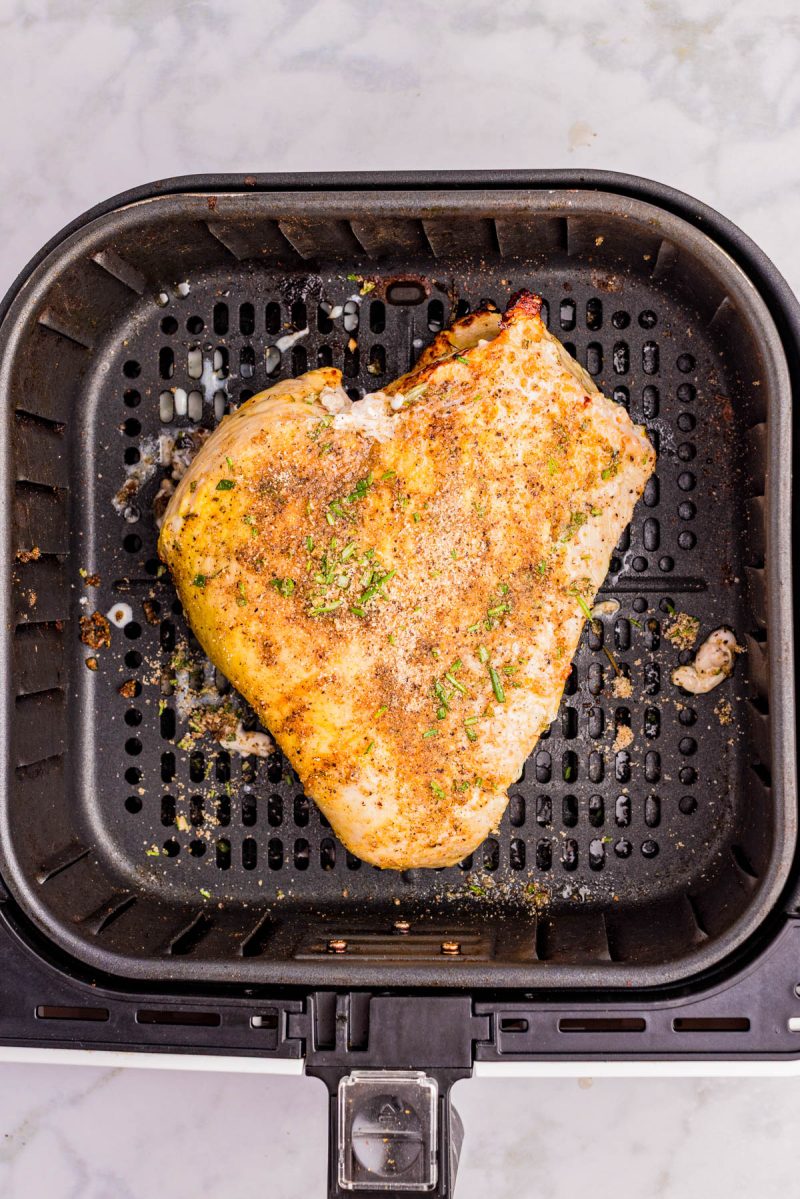 A seasoned chicken breast is cooking in an air fryer basket on a marble countertop.