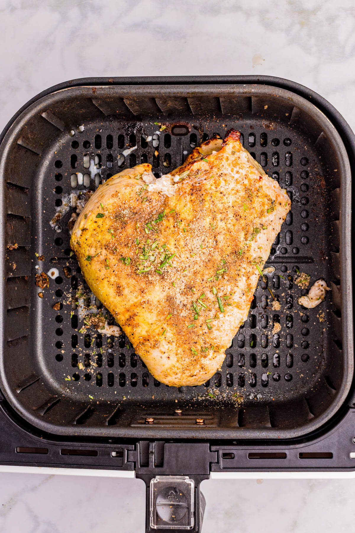 A seasoned chicken breast is cooking in an air fryer basket on a marble countertop.