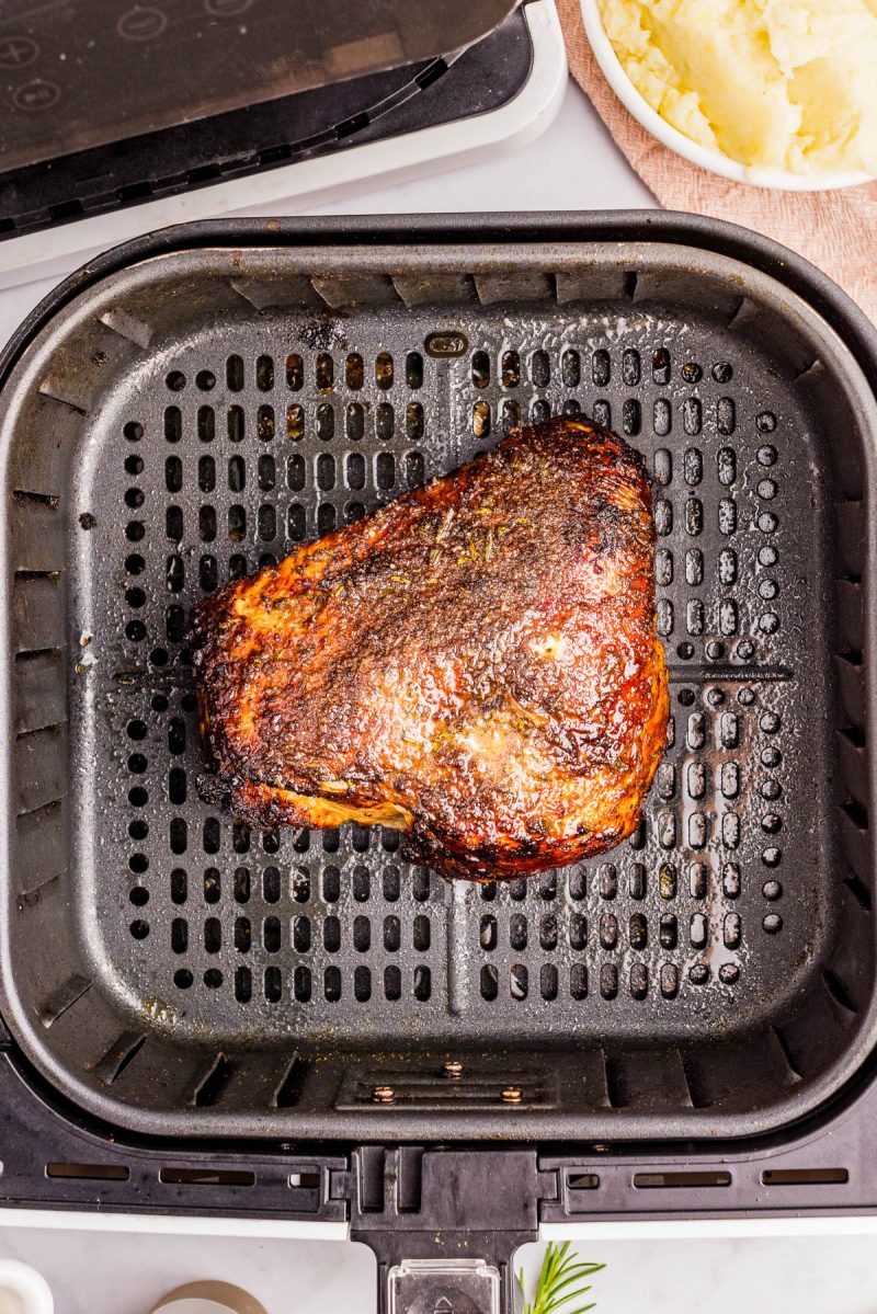 A cooked, seasoned beef roast sits in the basket of an air fryer. Mashed potatoes are visible in a bowl at the top right corner of the image.