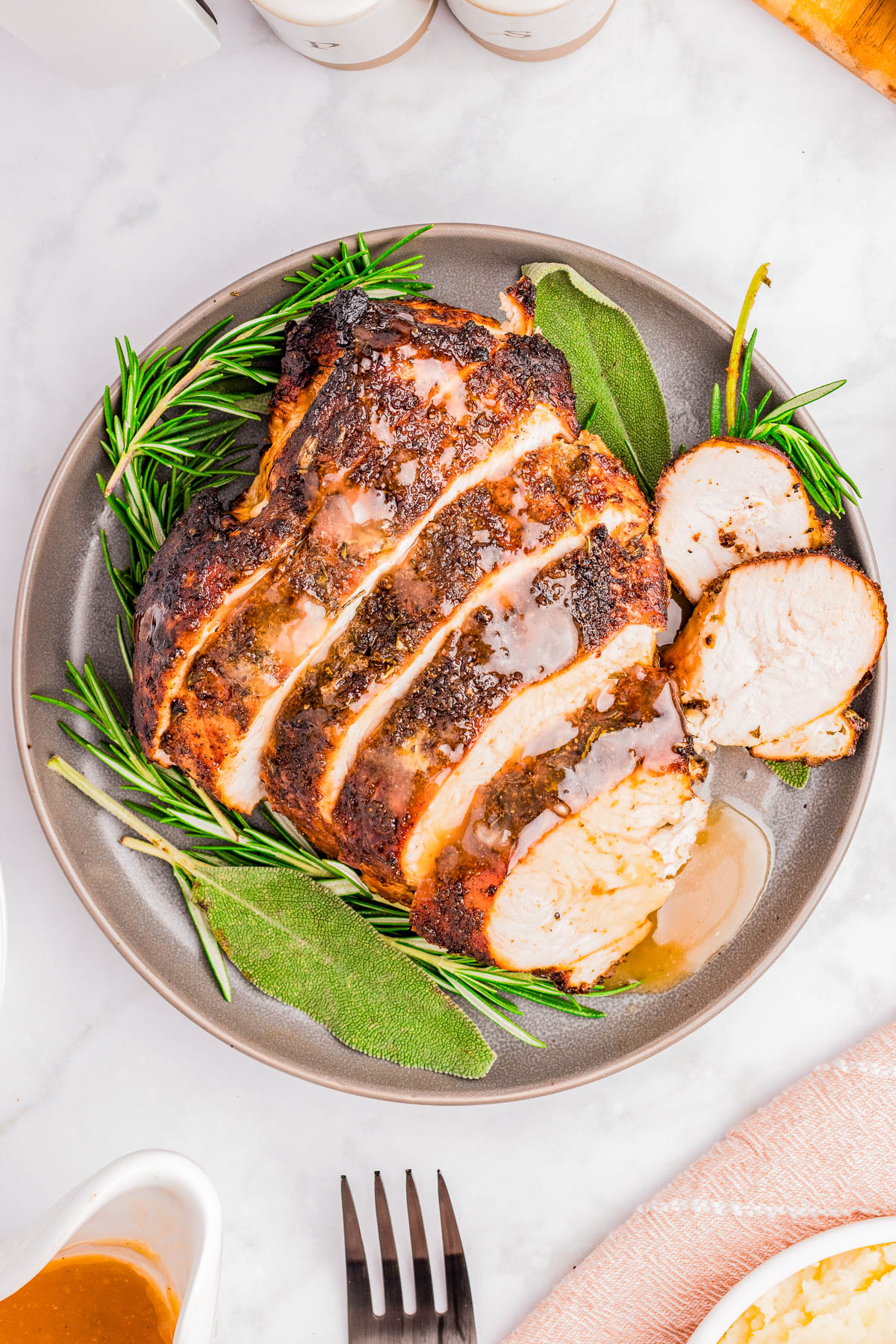 Sliced roasted turkey breast with herbs and gravy on a gray plate, garnished with fresh rosemary and sage, on a marble surface.