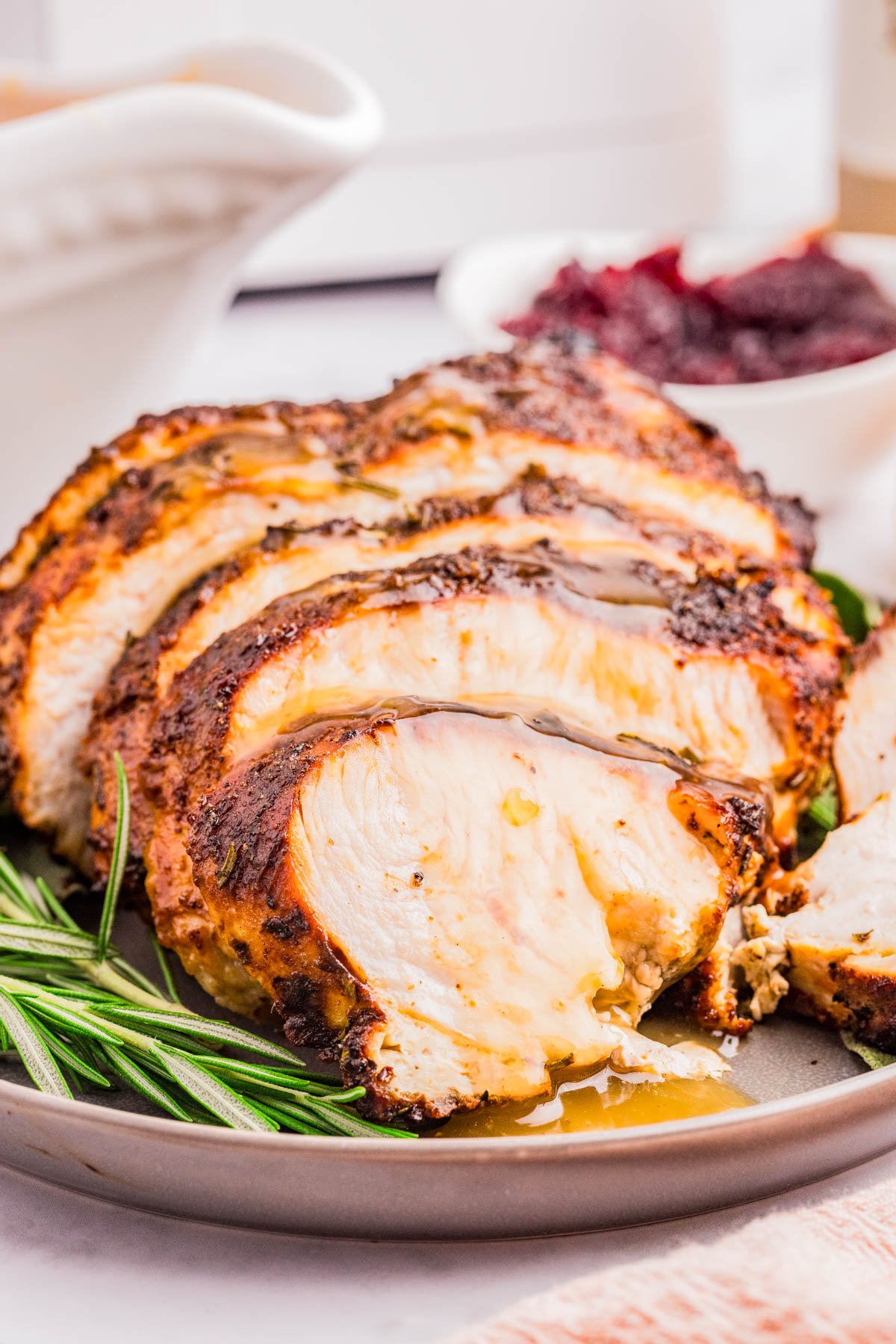 Sliced roasted turkey breast drizzled with gravy, served on a plate with fresh rosemary; a bowl of cranberry sauce is in the background.