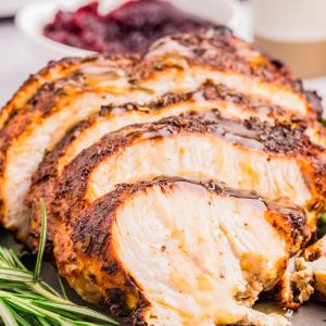 Sliced roasted turkey breast with herbs on a plate, garnished with rosemary, with a bowl of cranberry sauce in the background.