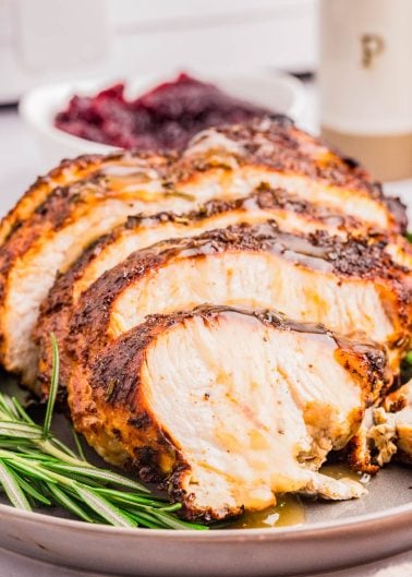 Sliced roasted turkey breast with herbs on a plate, garnished with rosemary, with a bowl of cranberry sauce in the background.