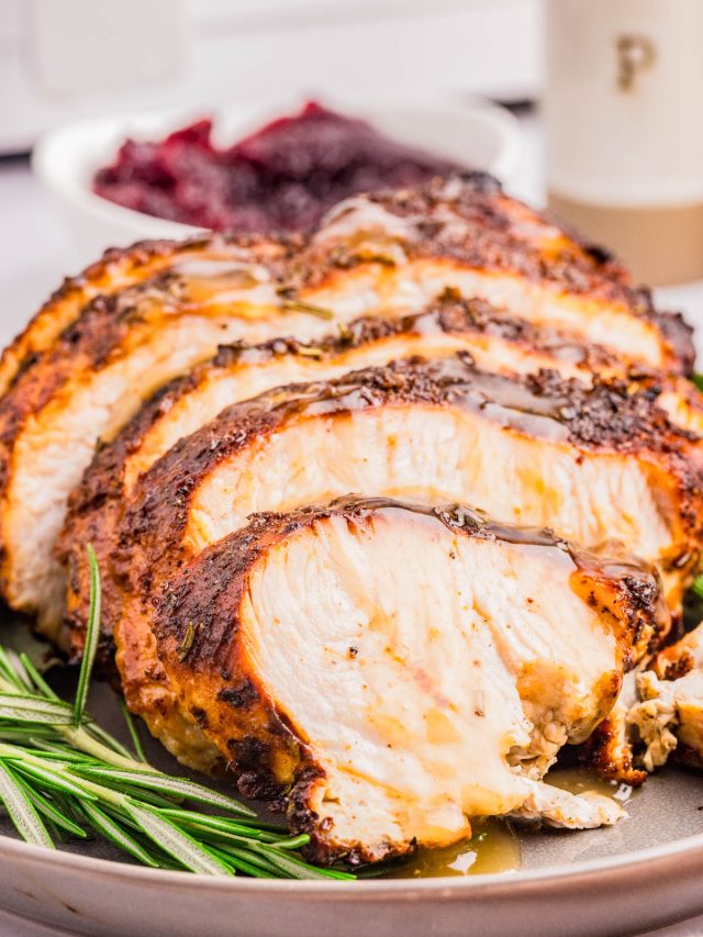 Sliced roasted turkey breast with herbs on a plate, garnished with rosemary, with a bowl of cranberry sauce in the background.