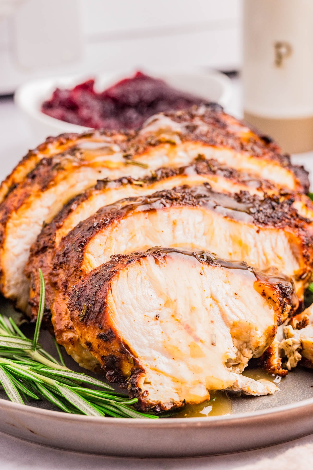 Sliced roasted turkey breast with herbs on a plate, garnished with rosemary, with a bowl of cranberry sauce in the background.