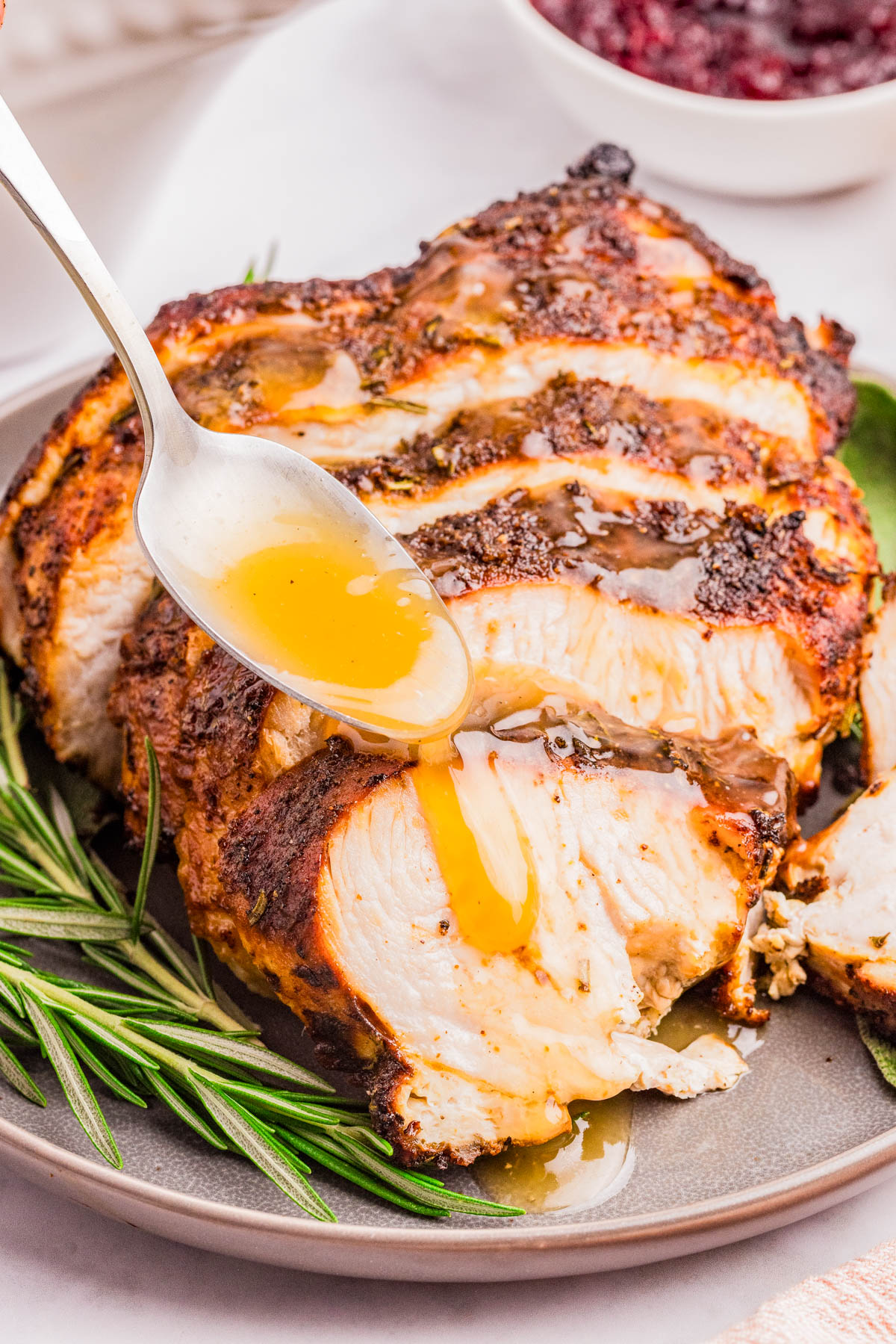 A sliced roasted turkey breast on a plate is being drizzled with gravy from a spoon, with a sprig of rosemary beside it.