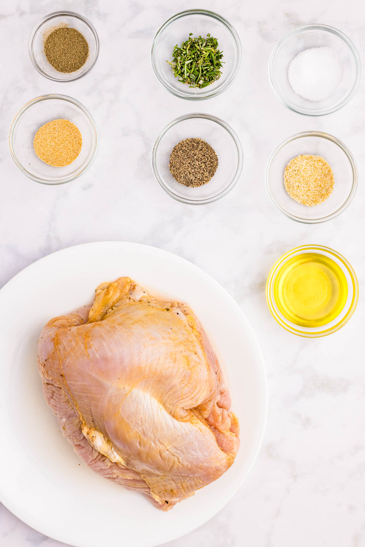 Raw turkey breast on a white plate surrounded by small bowls containing oil, salt, black pepper, dried herbs, garlic powder, onion powder, and ground pepper on a marble surface.