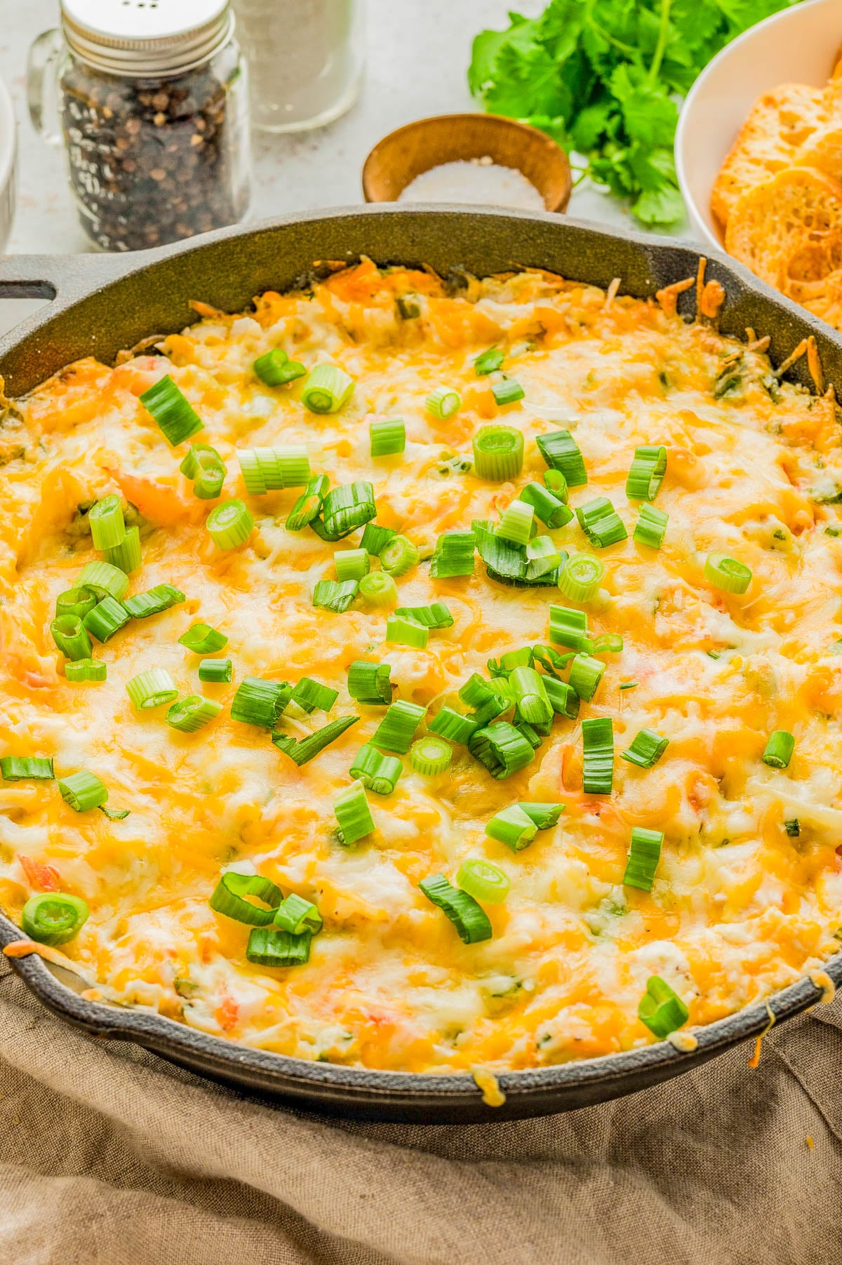 A skillet filled with baked cheesy dip, topped with chopped green onions, sits on a table with a jar of pepper, a bunch of cilantro, and a bowl of toasted bread slices nearby.