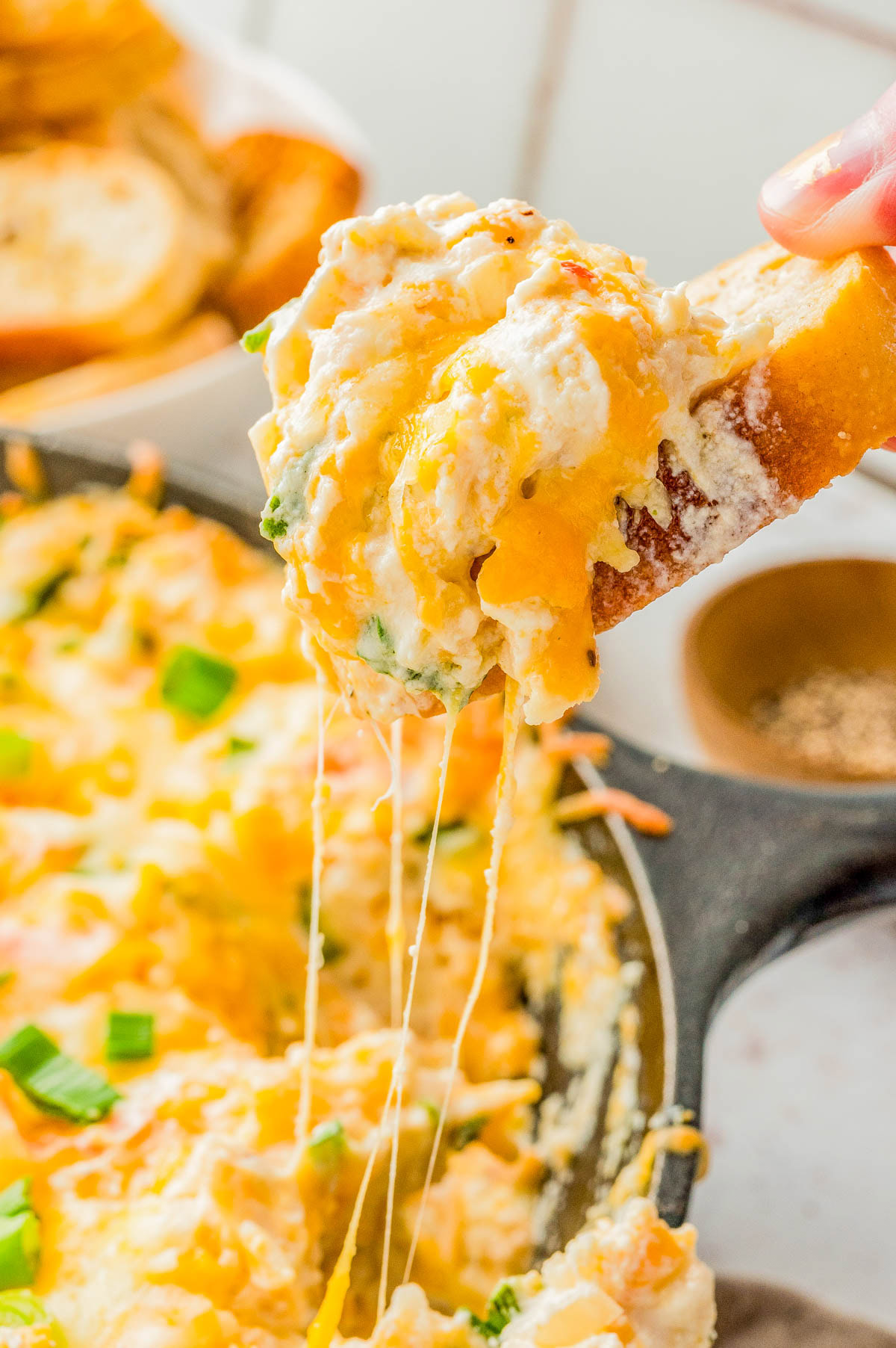 A hand dips a piece of bread into a cheesy, baked dip in a skillet, with melted cheese stretching between the bread and the dip.