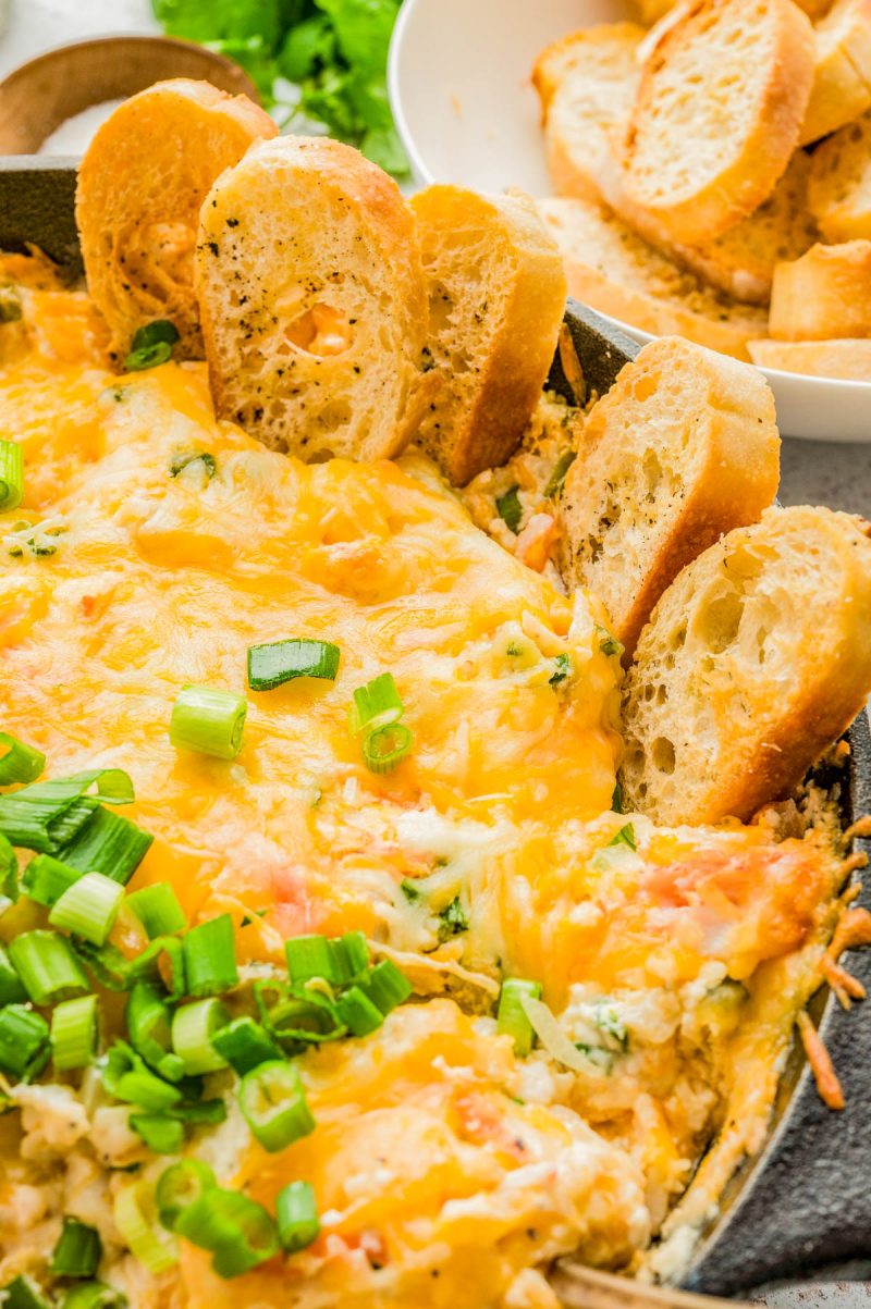 A skillet filled with cheesy baked dip, topped with chopped green onions and surrounded by toasted baguette slices, with a bowl of extra bread in the background.