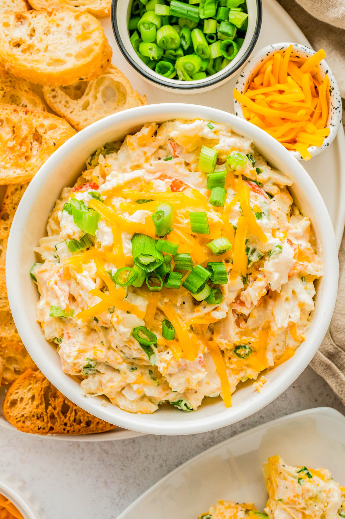 A bowl of creamy dip topped with shredded cheddar cheese and chopped green onions, surrounded by sliced bread, more cheese, and a small bowl of chopped green onions.