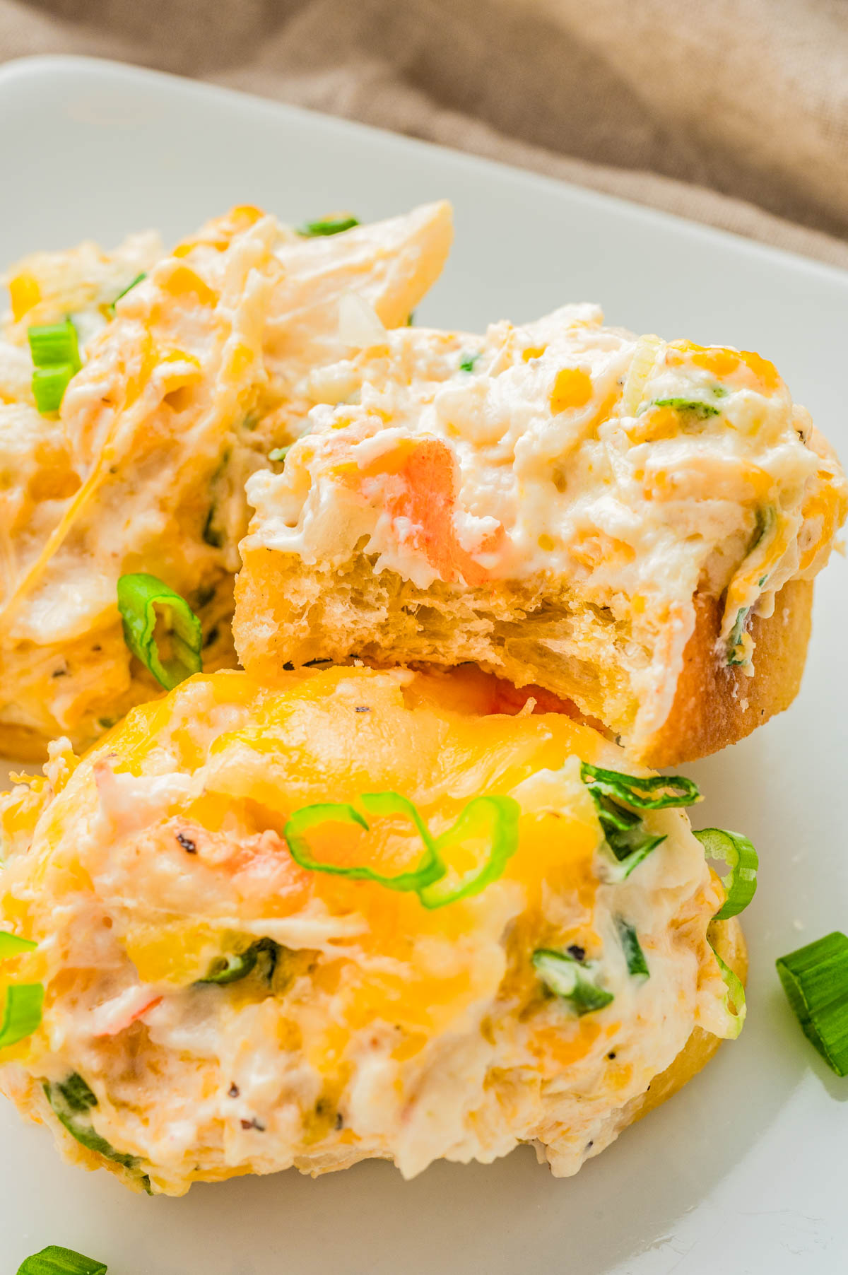 Two cheesy, baked crab dip appetizers garnished with chopped green onions, one with a bite taken out, on a white plate.