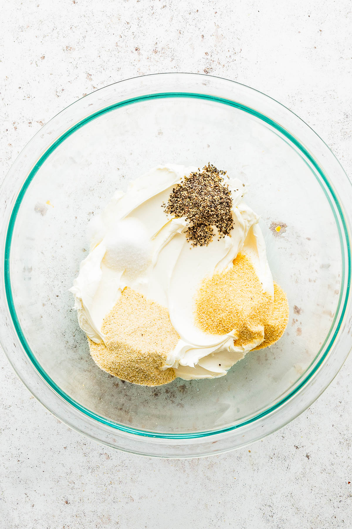 A glass bowl containing cream cheese, garlic powder, onion powder, salt, and ground black pepper on a white surface.