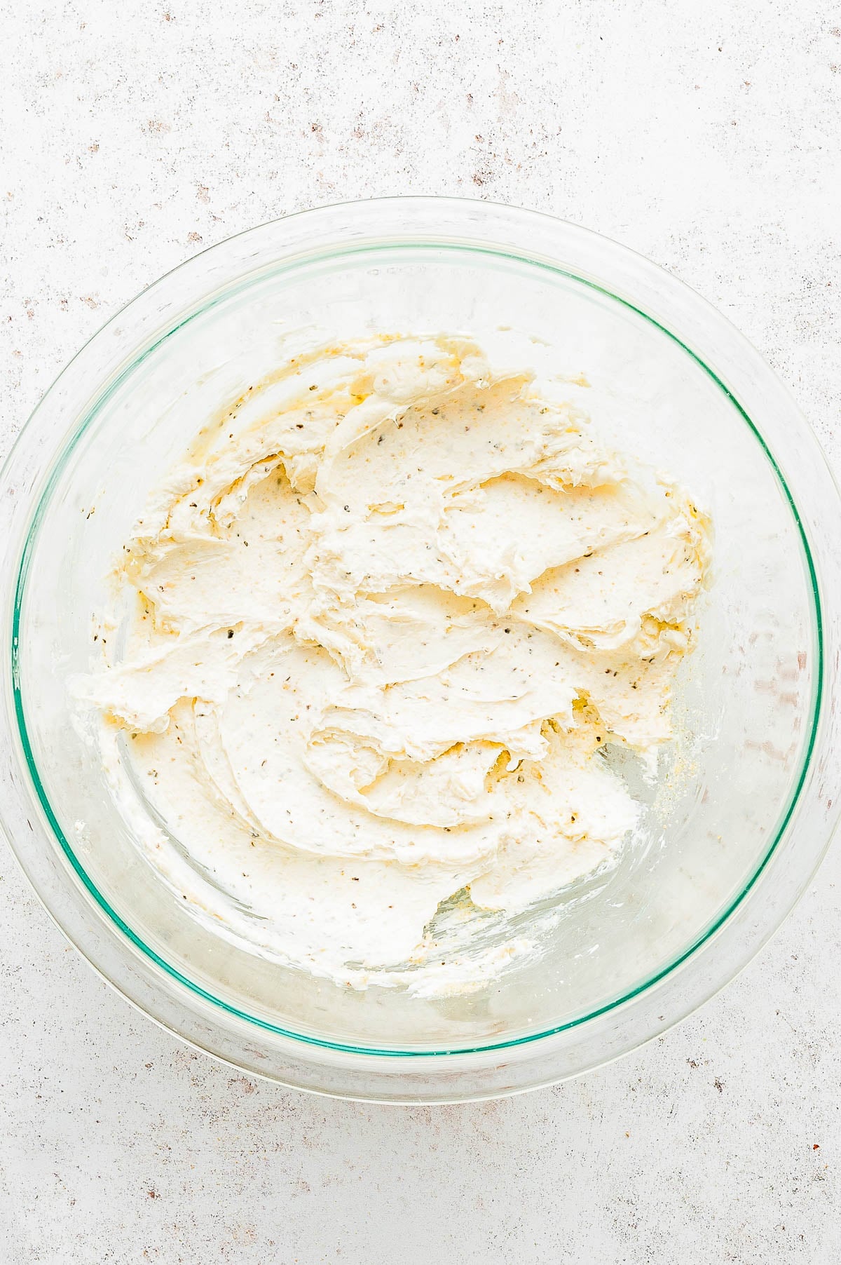 A clear glass bowl filled with a creamy, herb-flecked mixture on a light-colored, textured surface.