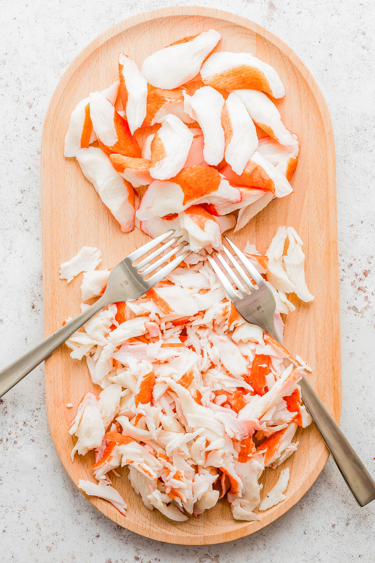 Two metal forks shredding imitation crab sticks on a wooden cutting board, with whole and shredded pieces separated.