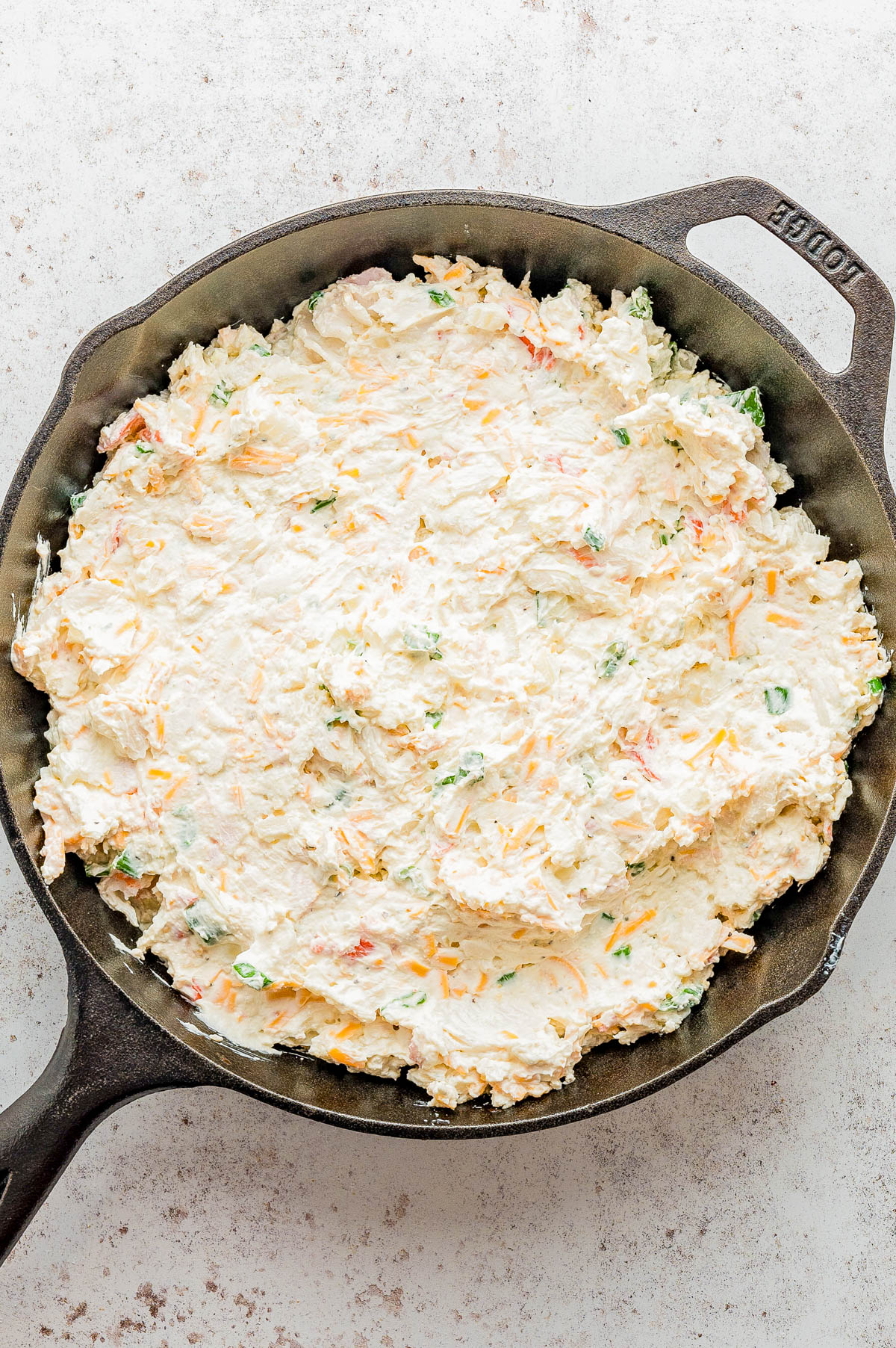 A cast iron skillet filled with an even layer of creamy dip mixed with shredded cheese and chopped vegetables on a light surface.