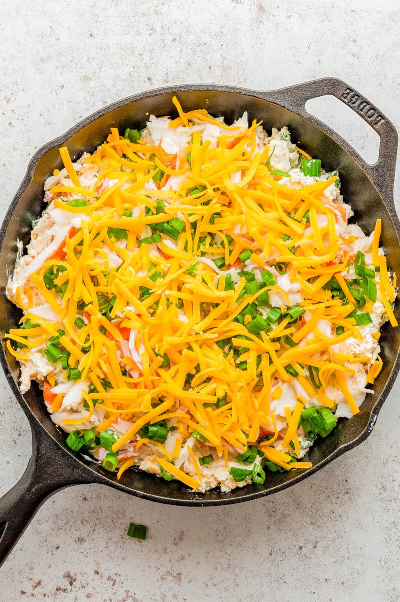 A cast iron skillet filled with a layered dip topped with shredded cheddar cheese, chopped green onions, and other ingredients on a light surface.