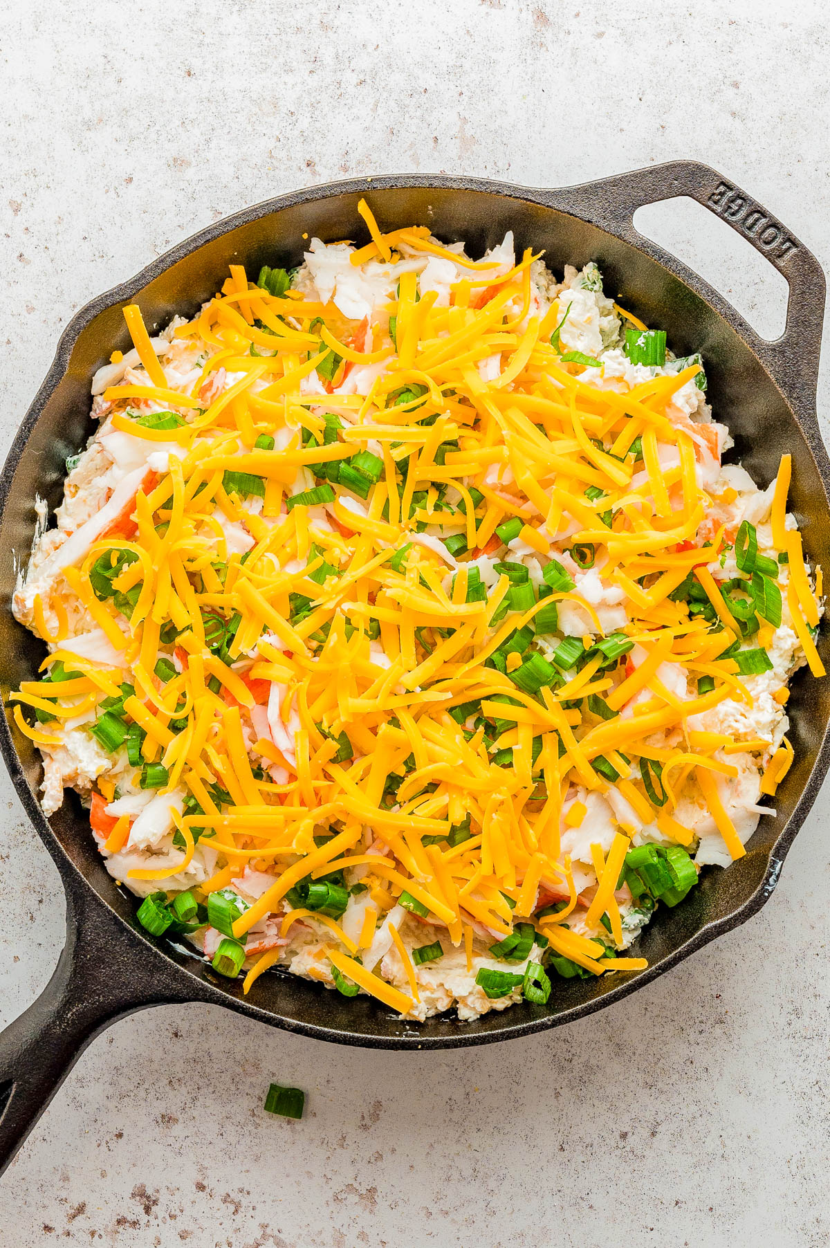 A cast iron skillet filled with a layered dip topped with shredded cheddar cheese, chopped green onions, and other ingredients on a light surface.