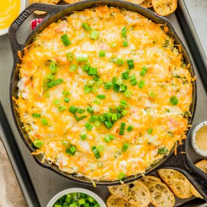 Cast iron skillet filled with baked cheesy dip topped with chopped green onions, surrounded by toasted baguette slices and bowls of chopped green onions.