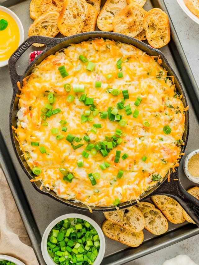 Cast iron skillet filled with baked cheesy dip topped with chopped green onions, surrounded by toasted baguette slices and bowls of chopped green onions.