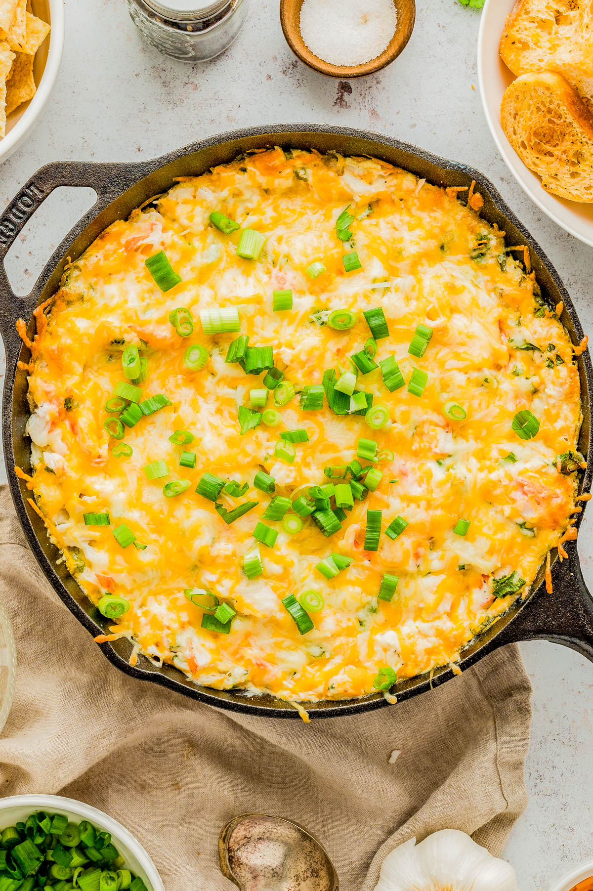 A cast iron skillet filled with a baked cheesy casserole topped with chopped green onions, surrounded by bowls of ingredients on a light surface.