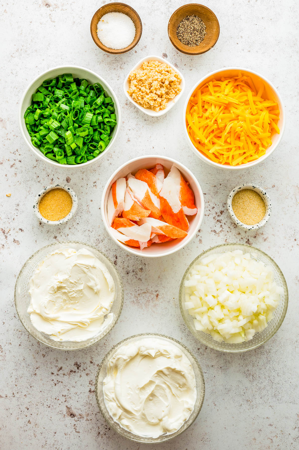 Ingredients for a recipe arranged in bowls, including chopped green onions, shredded cheddar cheese, imitation crab, cream cheese, mayonnaise, diced onions, garlic, salt, pepper, and seasonings.