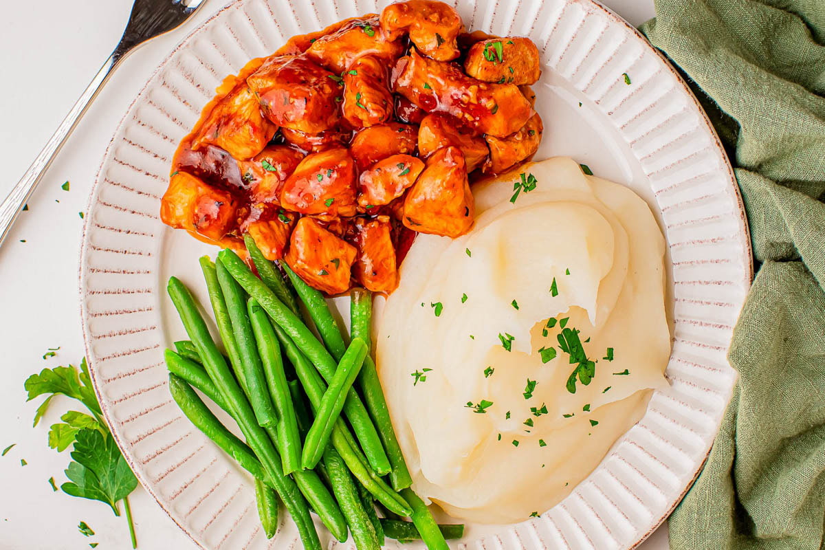 A plate with barbecue chicken pieces, mashed potatoes topped with parsley, and a serving of green beans.