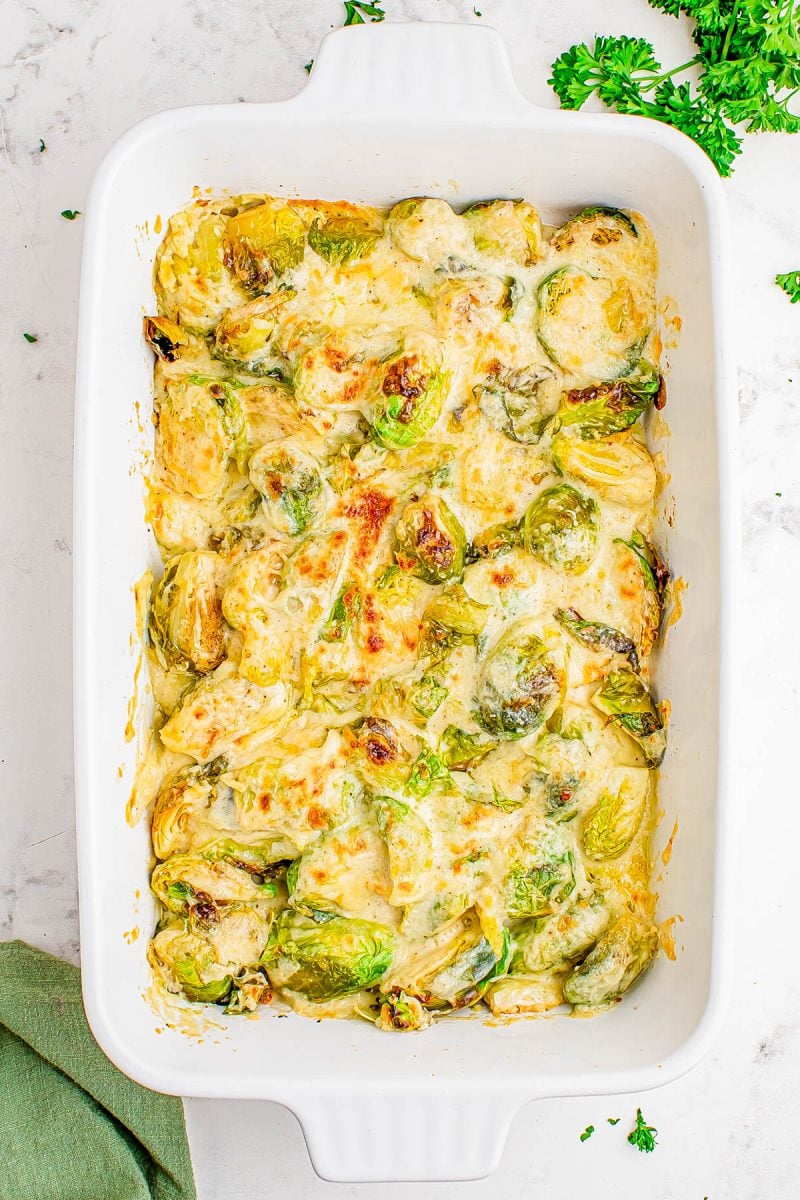 A white baking dish filled with baked Brussels sprouts topped with melted cheese, on a white surface with parsley garnish nearby.