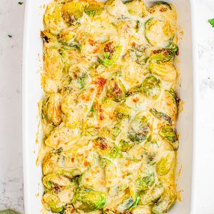 Baked Brussels sprouts covered in melted cheese sauce in a white rectangular casserole dish, with some herbs and kitchen items around it.