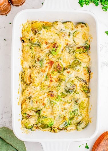 Baked Brussels sprouts covered in melted cheese sauce in a white rectangular casserole dish, with some herbs and kitchen items around it.