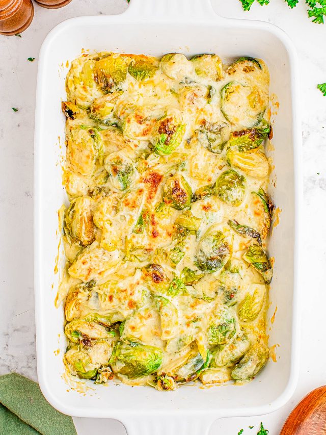 Baked Brussels sprouts covered in melted cheese sauce in a white rectangular casserole dish, with some herbs and kitchen items around it.