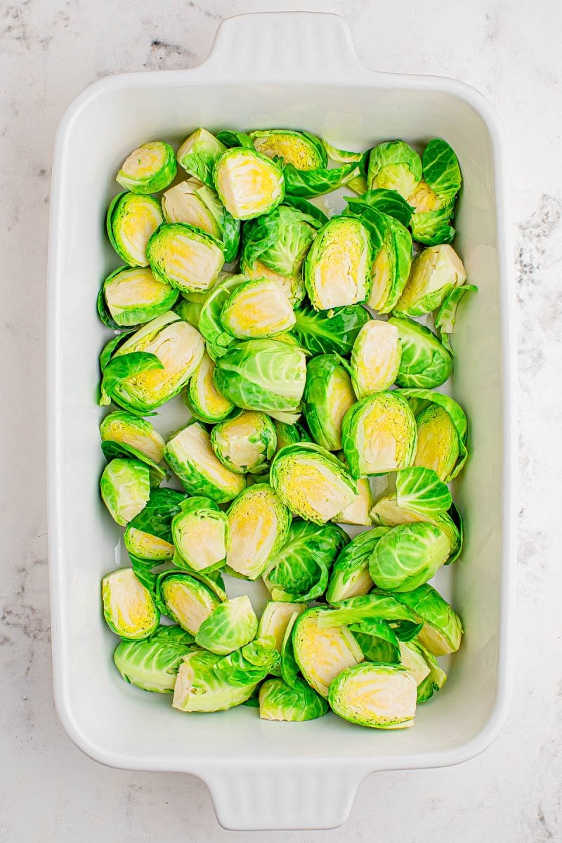 Halved Brussels sprouts arranged evenly in a white rectangular baking dish on a light marble surface.