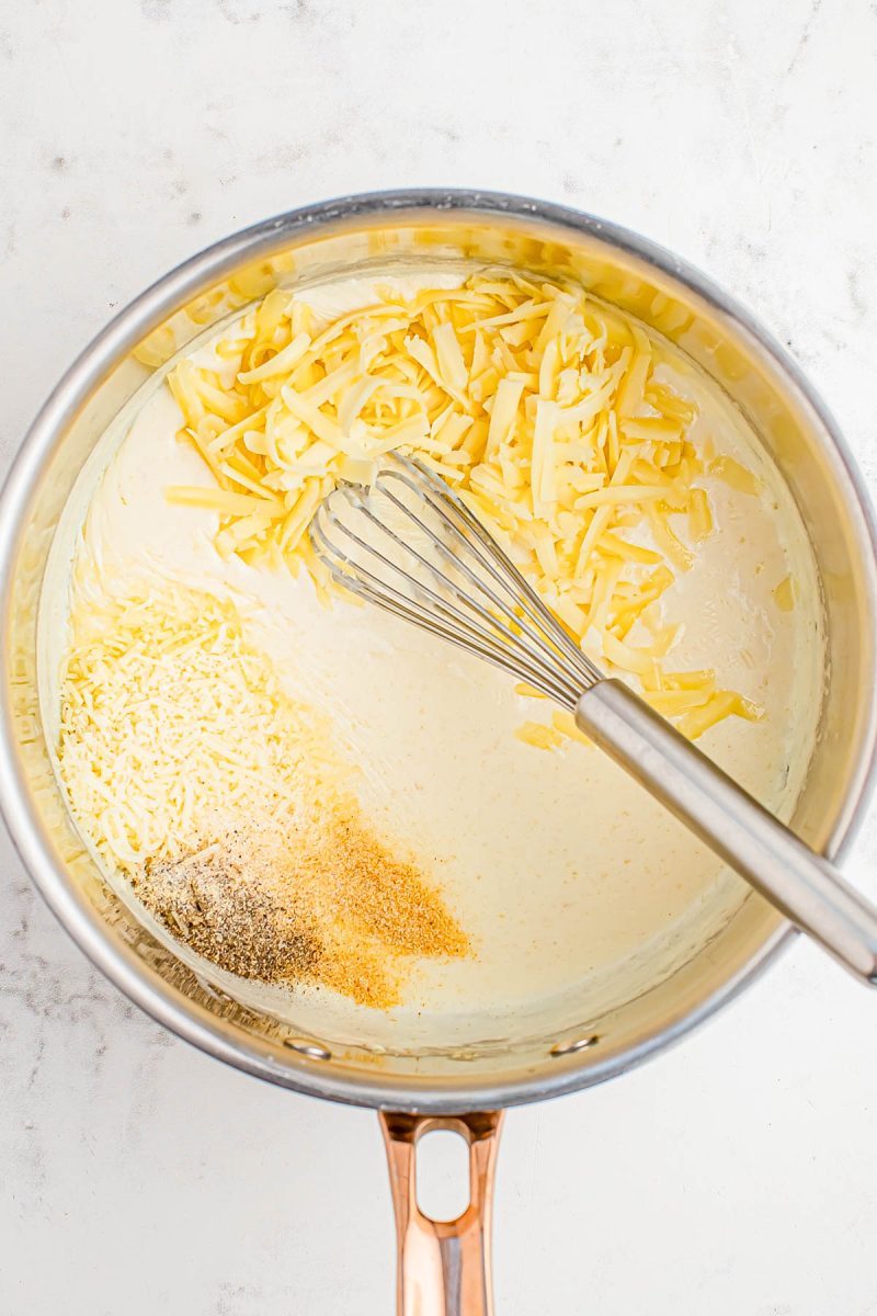A saucepan with shredded cheese, grated parmesan, ground black pepper, and garlic powder being mixed into a creamy sauce with a whisk.