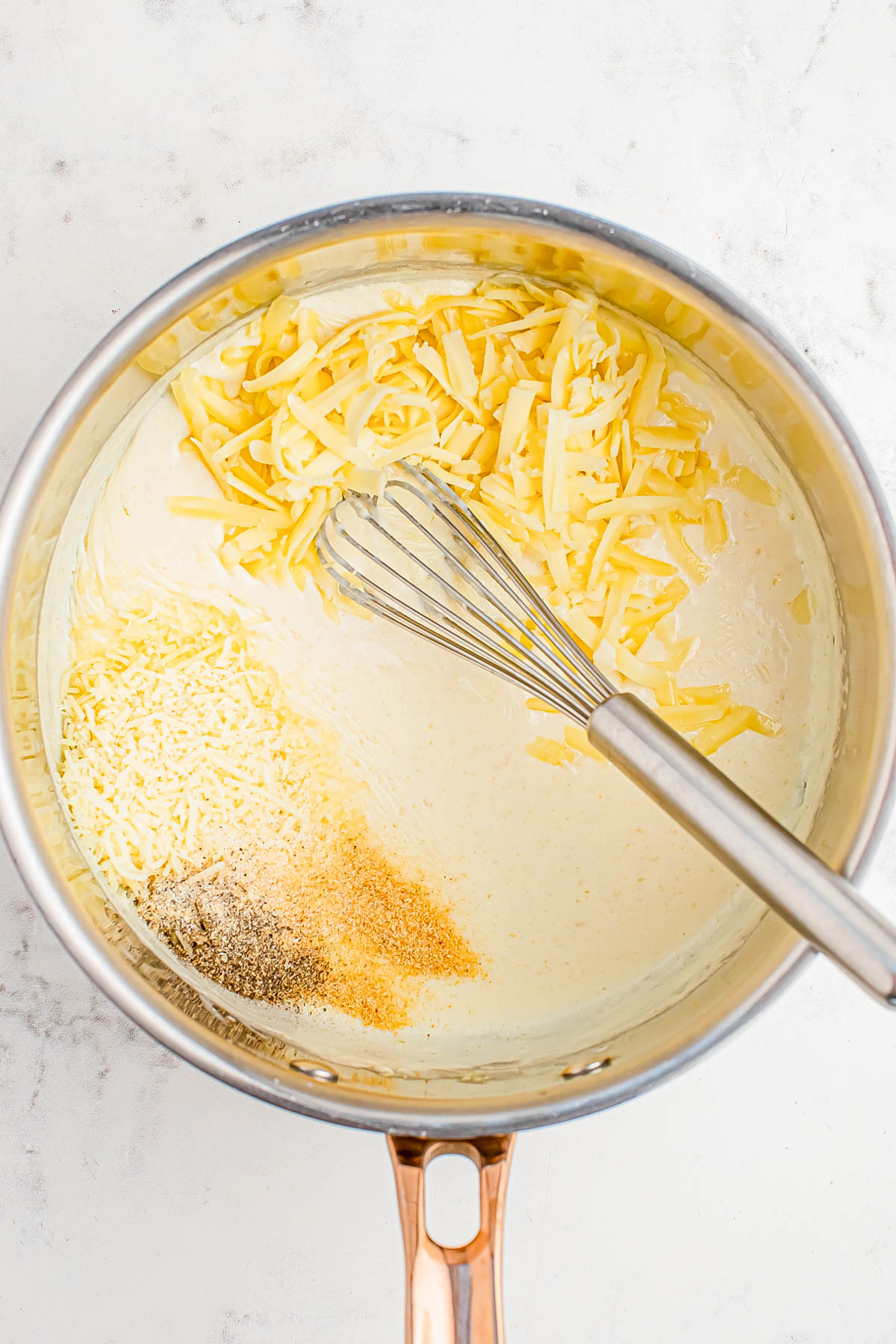 A saucepan with shredded cheese, grated parmesan, ground black pepper, and garlic powder being mixed into a creamy sauce with a whisk.
