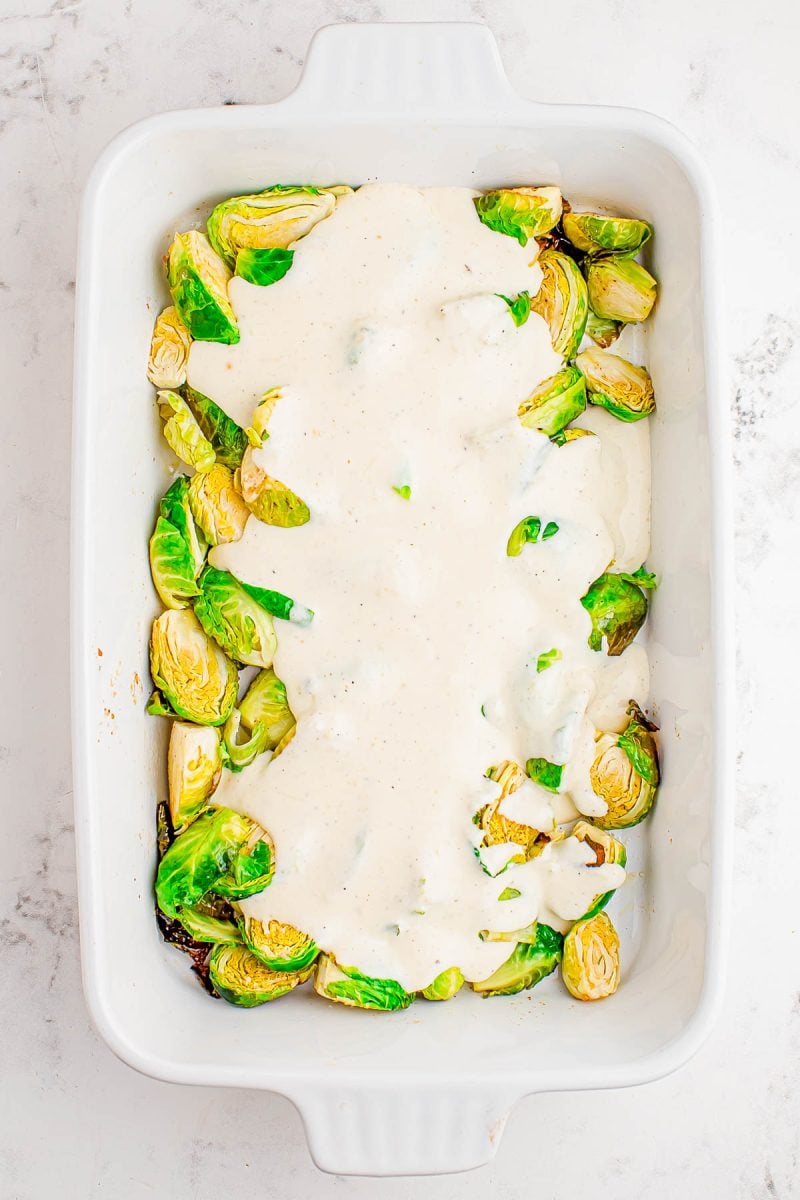 Halved Brussels sprouts in a white baking dish, partially covered with a creamy white sauce.