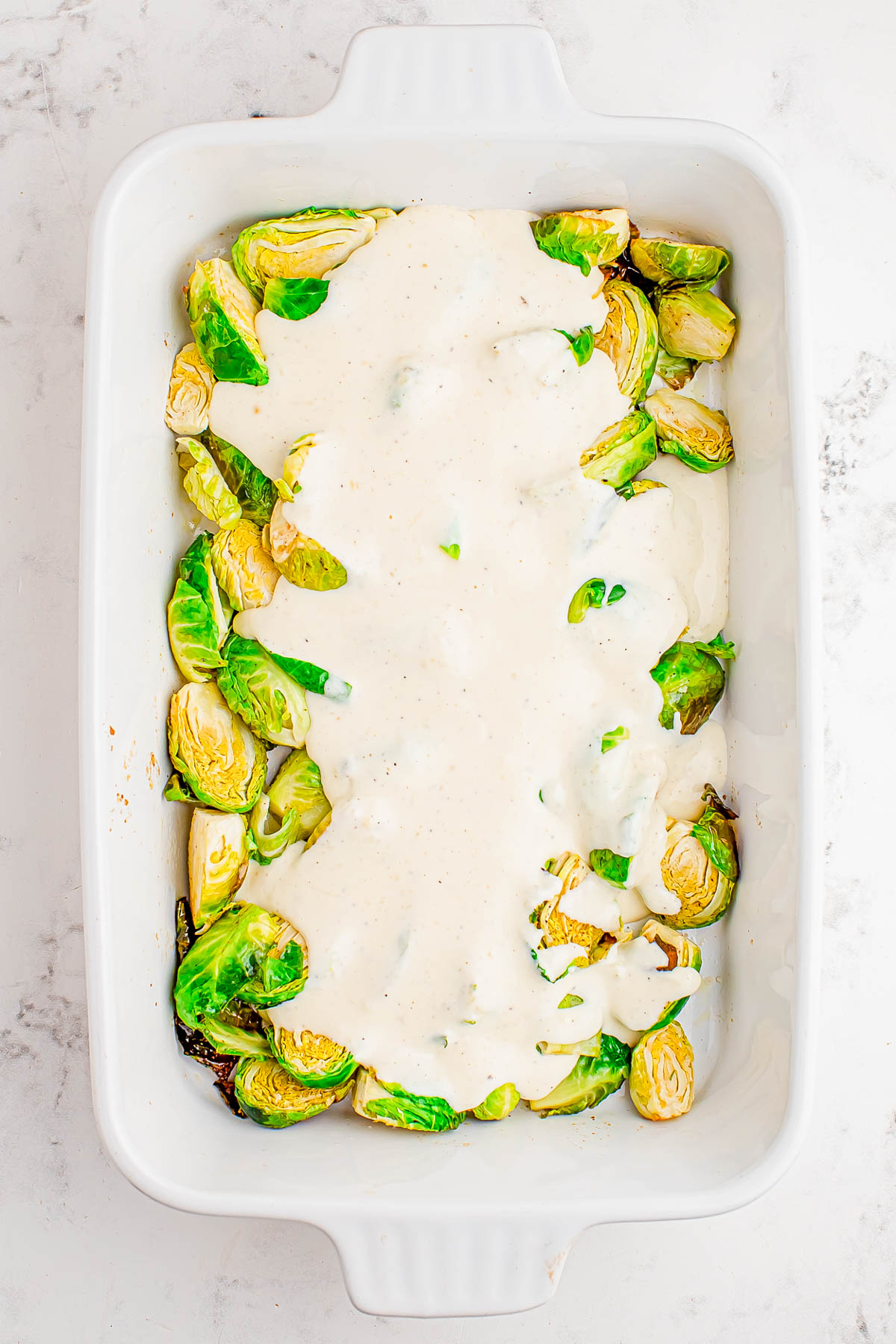 Halved Brussels sprouts in a white baking dish, partially covered with a creamy white sauce.