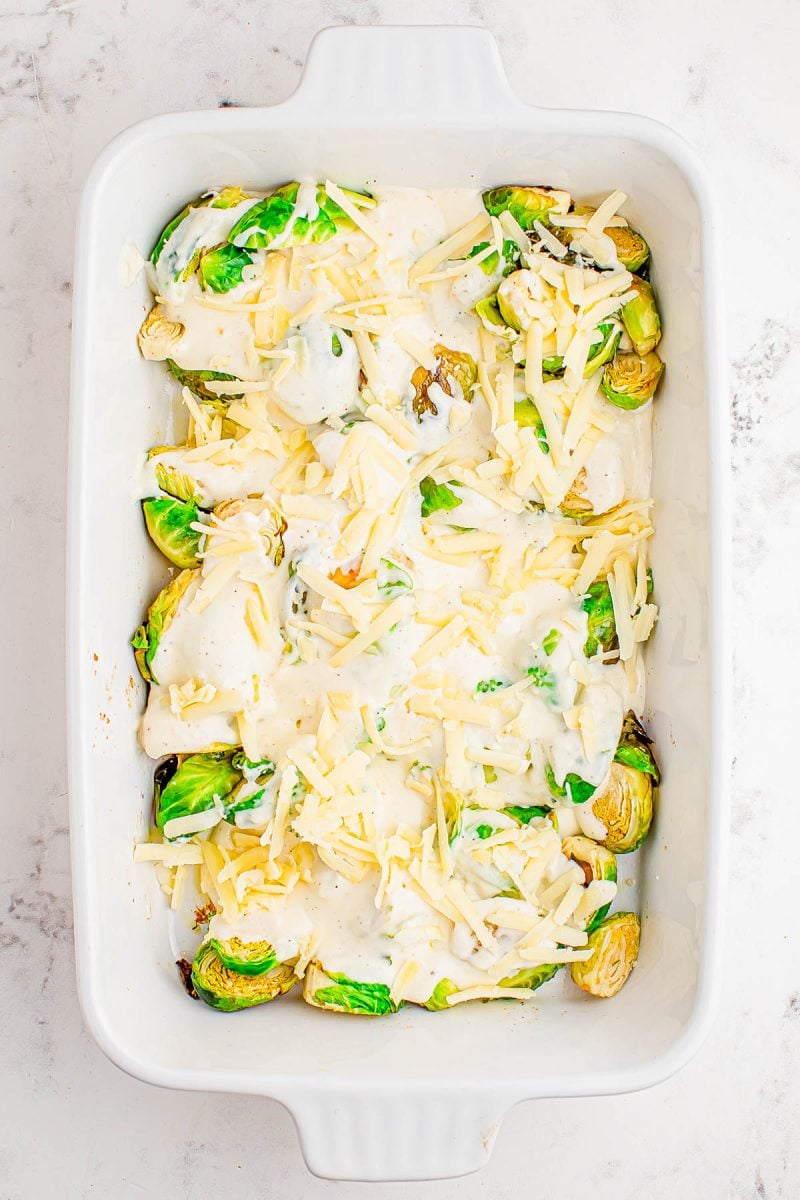 A white baking dish filled with halved Brussels sprouts topped with a creamy white sauce and shredded cheese, ready to be baked.