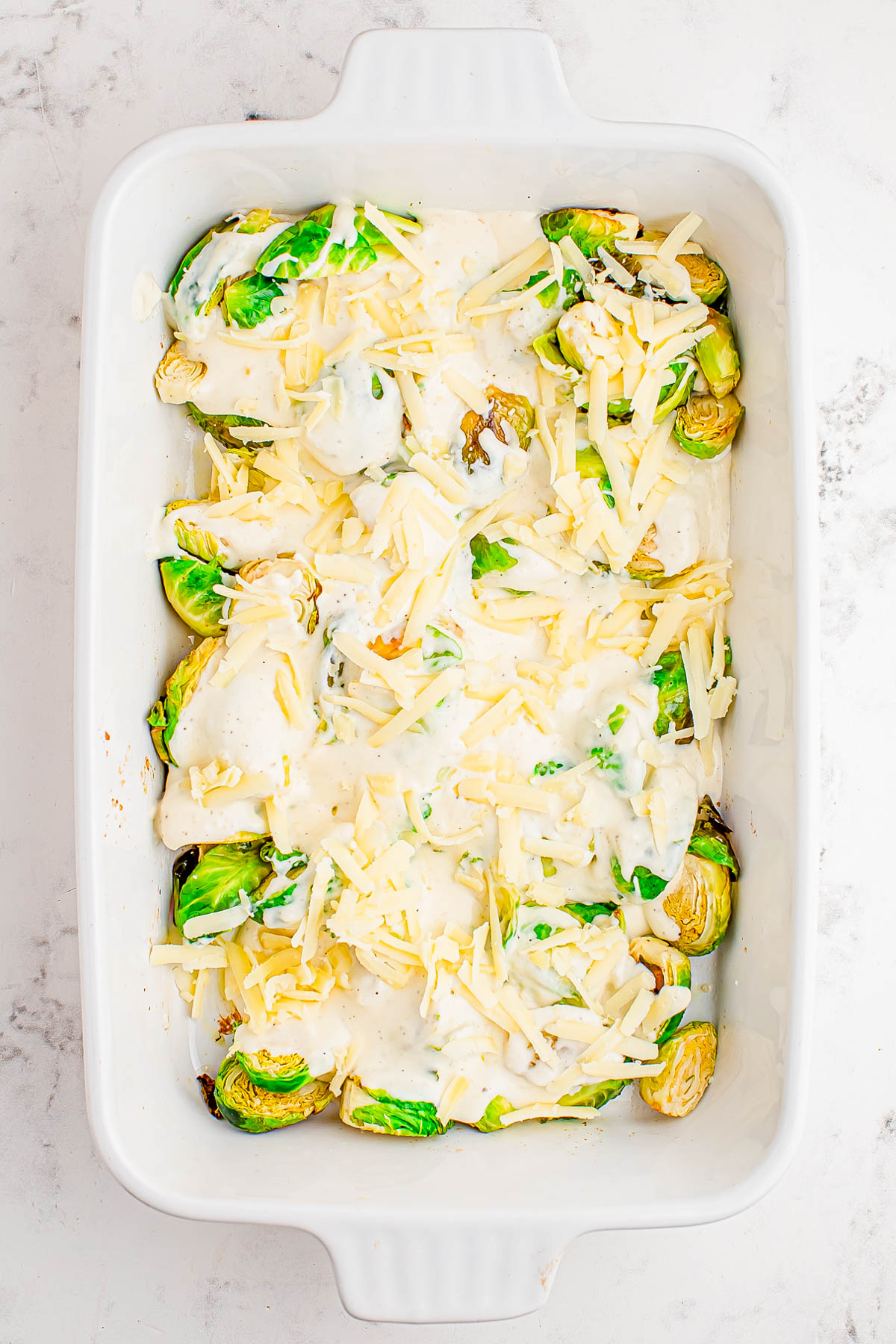 A white baking dish filled with halved Brussels sprouts topped with a creamy white sauce and shredded cheese, ready to be baked.