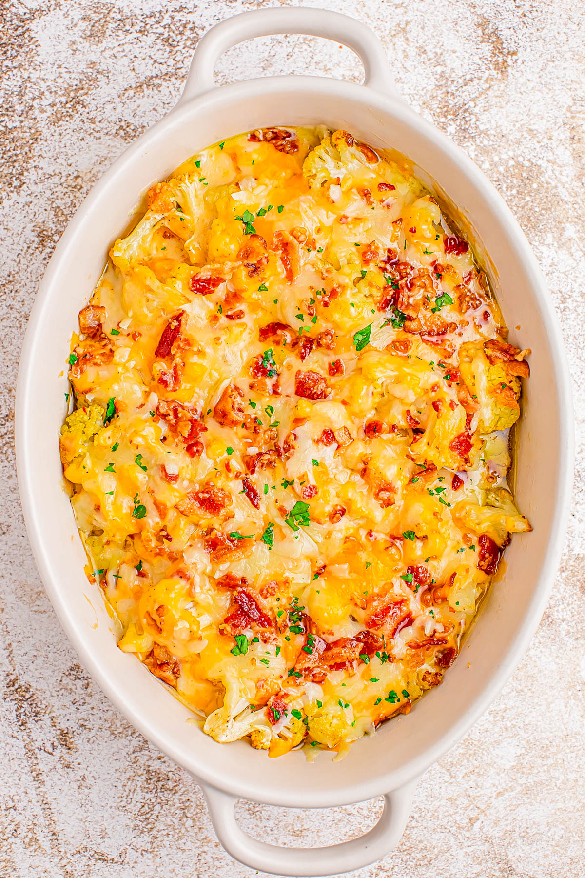 Oval baking dish filled with a cheesy cauliflower casserole topped with chopped bacon and garnished with parsley.