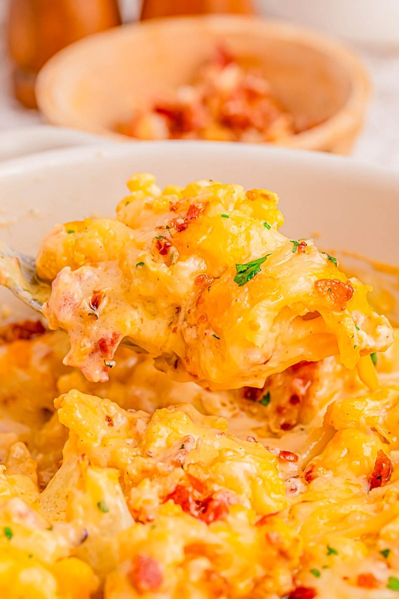 A close-up of a cheesy, baked pasta dish with visible bacon bits and herbs, being scooped from a white bowl with a spoon.